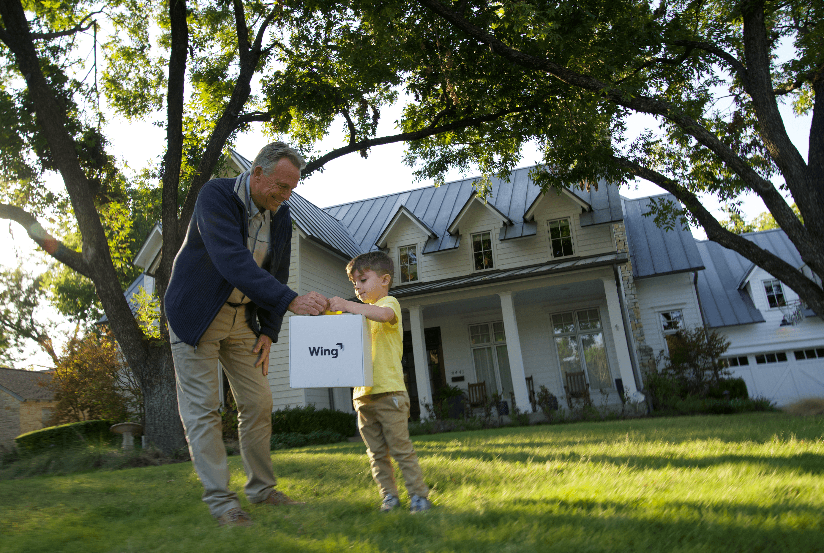 A grandfather and child stand on a lush green lawn in front of a large, charming house with tall trees, as they receive a drone delivery package marked "Wing".